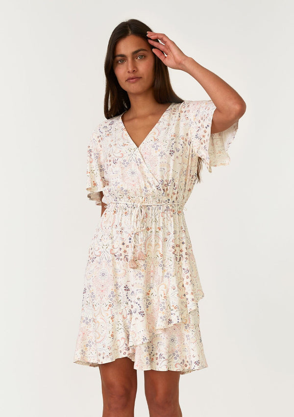 [Color: Natural/Peach] A front facing image of a brunette model wearing a bohemian ivory and pink floral print mini dress. With short flutter sleeves, a surplice v neckline, an elastic waist, a tassel tie waist belt, and a ruffled hemline. 