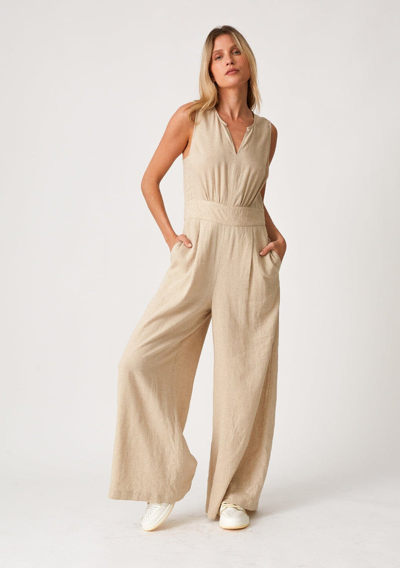 [Color: Wheat] A front facing image of a blonde model wearing linen blend full length jumpsuit. With a wide leg, side pockets, a v neckline, and a back zip closure.