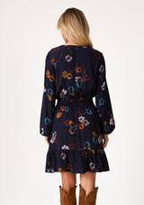 [Color: Navy/Mustard] A back facing image of a blonde model wearing a bohemian fall mini dress in a blue floral print. With long sleeves, a v neckline, a smocked elastic waist, a drawstring waist with tassel ties, a tiered mini skirt, and a self covered button front top. 