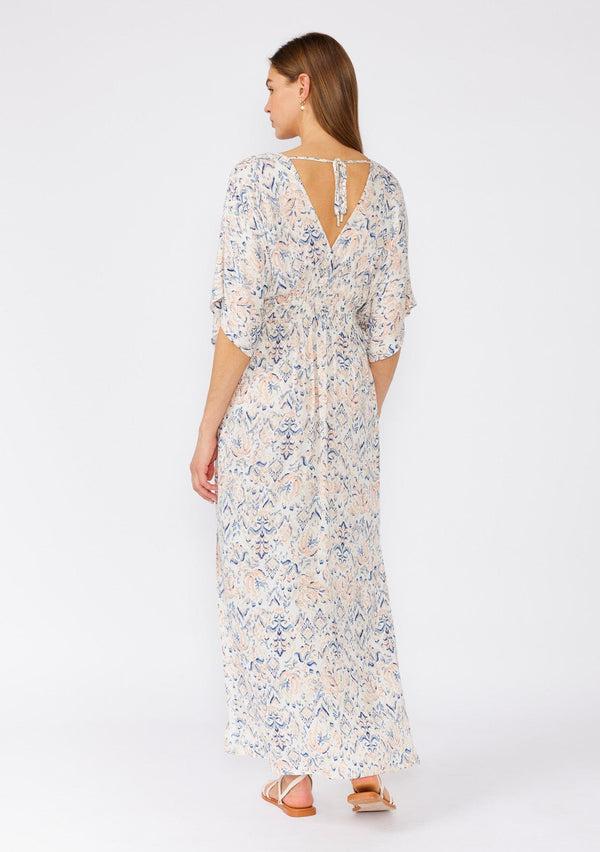 [Color: Cream/Dusty Blue] A back facing image of a brunette model wearing a flowy bohemian beach dress in a cream and blue abstract print. With flowy half length sleeves, a v neckline in the front and back, a smocked elastic waist, side slits, and an open back with tie detail. 