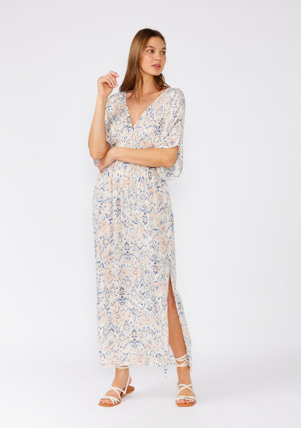 [Color: Cream/Dusty Blue] A front facing image of a brunette model wearing a flowy bohemian beach dress in a cream and blue abstract print. With flowy half length sleeves, a v neckline in the front and back, a smocked elastic waist, side slits, and an open back with tie detail. 