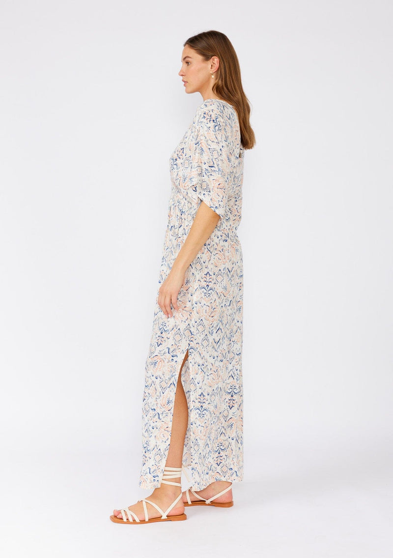 [Color: Cream/Dusty Blue] A side facing image of a brunette model wearing a flowy bohemian beach dress in a cream and blue abstract print. With flowy half length sleeves, a v neckline in the front and back, a smocked elastic waist, side slits, and an open back with tie detail. 