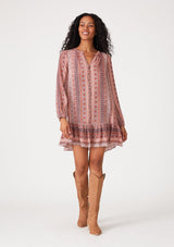 [Color: Cinnamon/Coral] A front facing image of a brunette model wearing a bohemian fall mini dress in a pink print. With long sleeves, a flowy baby doll silhouette, a tiered skirt, a self covered button front, and a v neckline. 