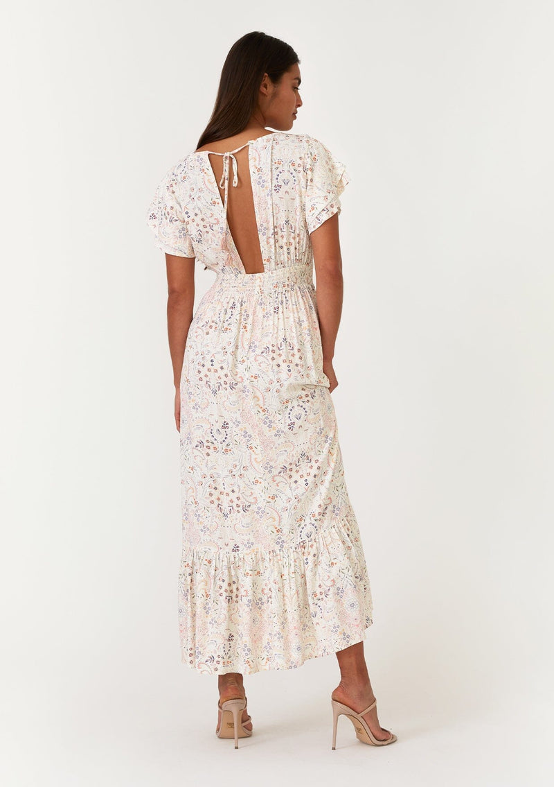 [Color: Natural/Peach] A back facing image of a brunette model wearing a bohemian special occasion maxi dress in a natural and pink floral print. With short flutter sleeves, a v neckline, a tiered skirt, an open back with tassel tie detail, and a half smocked elastic waist at the back. 