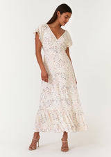 [Color: Natural/Peach] A front facing image of a brunette model wearing a bohemian special occasion maxi dress in a natural and pink floral print. With short flutter sleeves, a v neckline, a tiered skirt, an open back with tassel tie detail, and a half smocked elastic waist at the back. 