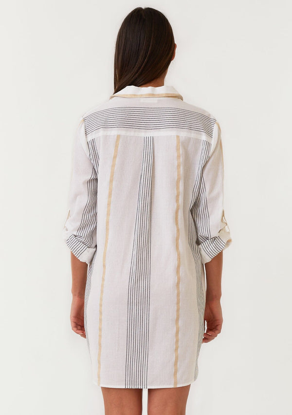 [Color: White/Gold] A back facing image of a brunette model wearing a white, black and gold striped mini shirt dress. A cotton spring dress with textured embroidered stripe details, a gold metallic lurex stripe, long rolled sleeves with a button tab closure, a split v neckline, a collared neckline, and side pockets. 