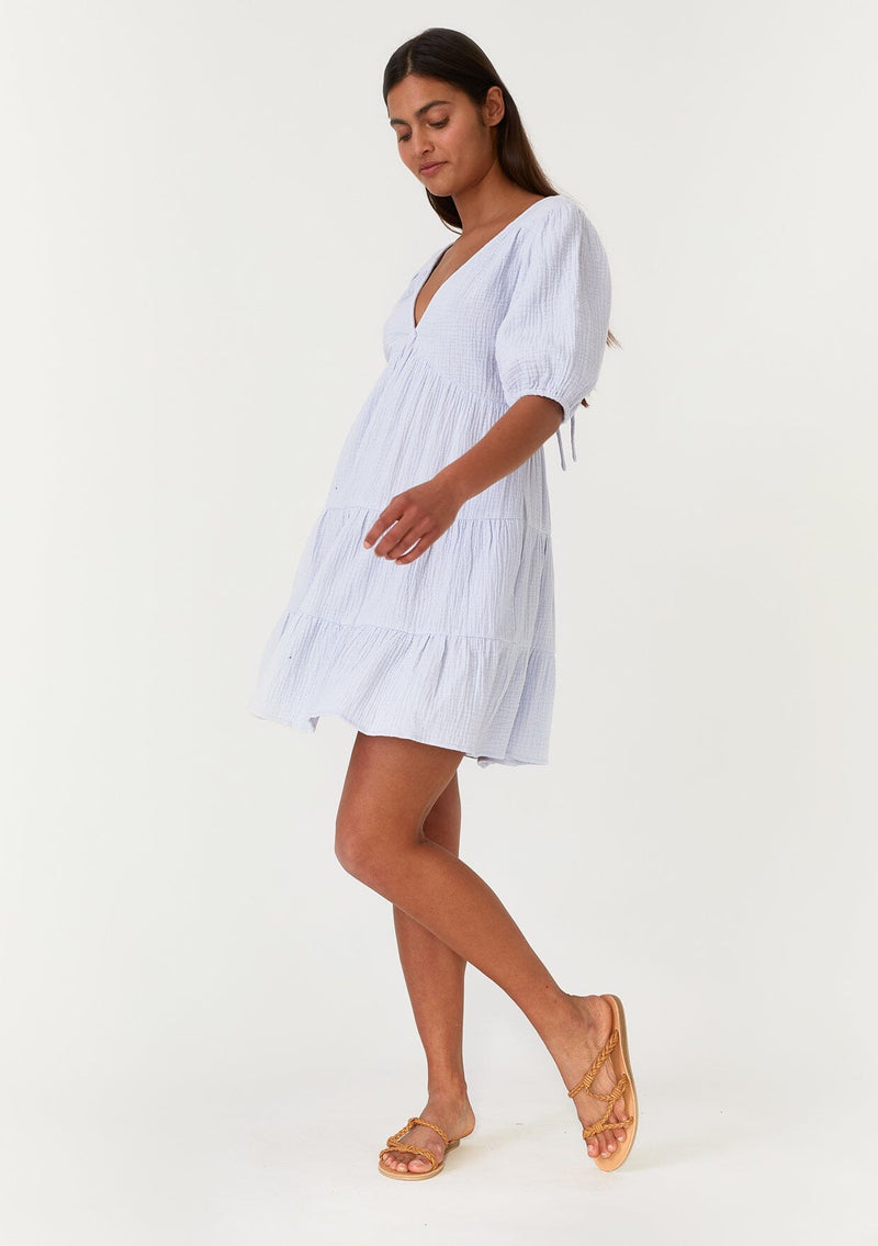 [Color: Dusty Blue] A full body side facing image of a brunette model wearing a cute bohemian spring mini dress crafted in a soft cotton gauze. With short puff sleeves, a v neckline in the front and back, an empire waist, a tiered skirt, and an open back detail with a tie closure. 