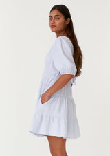 [Color: Dusty Blue] A side facing image of a brunette model wearing a cute bohemian spring mini dress crafted in a soft cotton gauze. With short puff sleeves, a v neckline in the front and back, an empire waist, a tiered skirt, and an open back detail with a tie closure. 