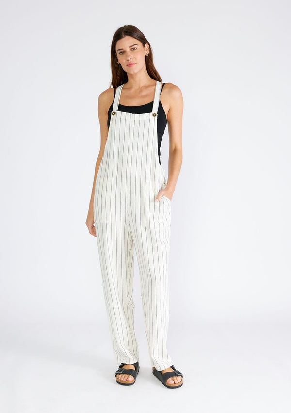 [Color: Natural/Black] A front facing image of a brunette model wearing a cool sleeveless jumpsuit crafted from a linen blend, in an ivory and black stripe. With a long straight leg, a racerback, tank top straps that button at the front, and side pockets. 