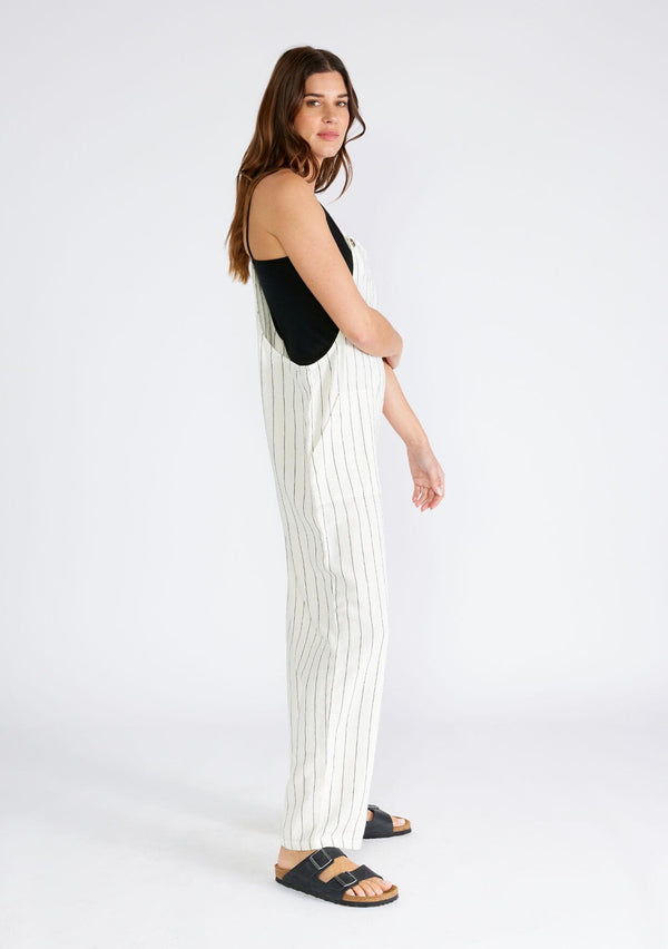 [Color: Natural/Black] A side facing image of a brunette model wearing a cool sleeveless jumpsuit crafted from a linen blend, in an ivory and black stripe. With a long straight leg, a racerback, tank top straps that button at the front, and side pockets. 