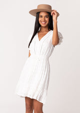 [Color: White] A front facing image of a brunette model wearing a white bohemian summer mini dress in embroidered eyelet. With short flutter sleeves, a smocked elastic waist, a v neckline, a tie waist accent, a tiered high low mini skirt, and lattice trim. 
