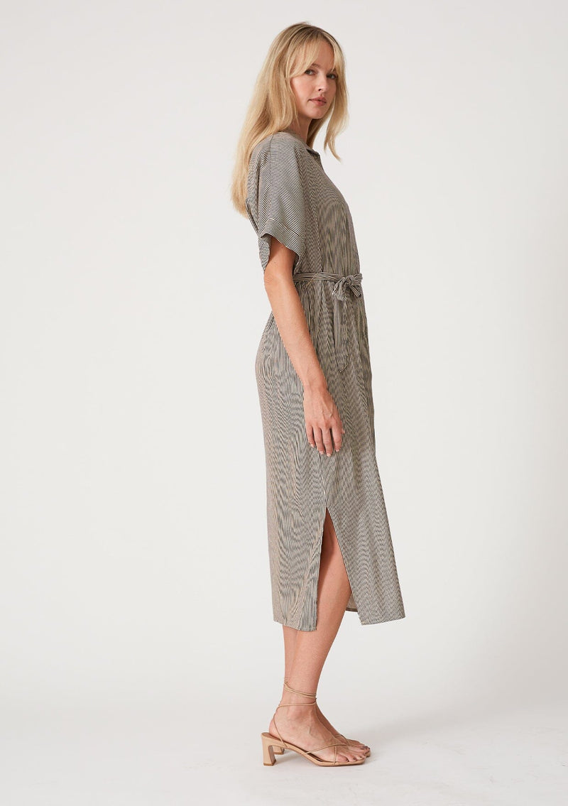 [Color: Taupe/Navy] A side facing image of a blonde model wearing a relaxed fit mid length shirt dress in a taupe and navy blue stripe. With short dolman sleeves, a collared neckline, a concealed button front, side pockets, a waist belt, and side slits. 