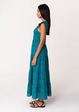 [Color: Teal] A side facing image of a brunette model wearing a bohemian cotton maxi dress in a teal embroidered eyelet. With a slim fit smocked bodice, a square neckline, short flutter sleeves, and a flowy long tiered skirt. 