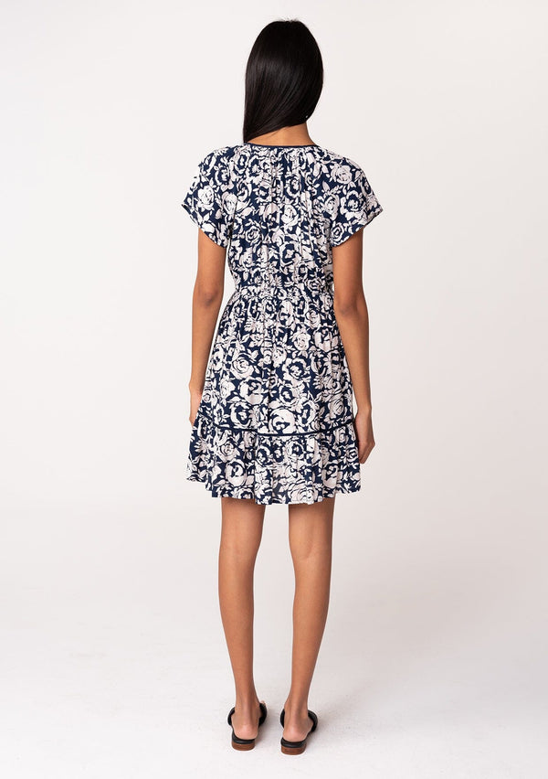 [Color: Navy/Natural] A back facing image of a brunette model wearing a navy blue floral print summer mini dress. With short flutter sleeves, a split v neckline with tassel ties, an elastic waist, a self covered button front top, and a tiered mini skirt. 