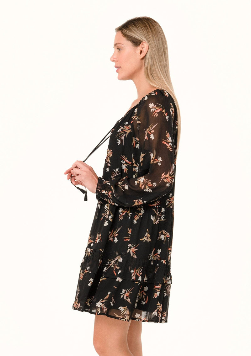 [Color: Black/Peach] A front facing image of a blonde model standing outside wearing a pretty bohemian chiffon mini dress in a black and peach floral print. With sheer long sleeves, ruffled elastic wrist cuffs, a tiered skirt, a v neckline with tassel ties, and a decorative self covered button front.