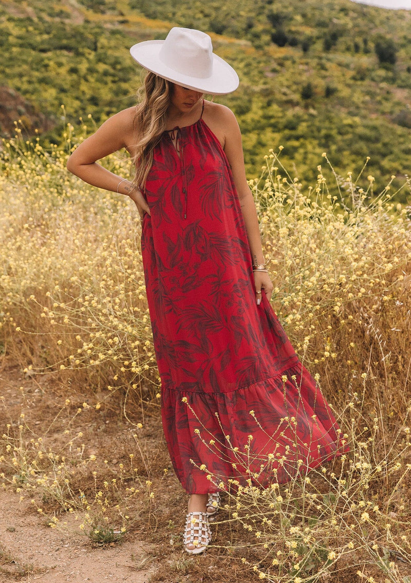 [Color: Wine/Charcoal] A front facing image of a blonde model standing outside wearing a dark red chiffon halter maxi dress. With a drawstring halter neckline, a front keyhole, a tiered skirt, and an ultra flowy silhouette.