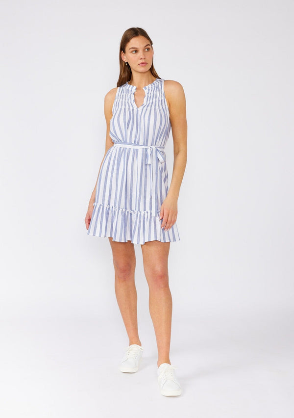 [Color: White/Blue] A full body front facing image of a brunette model wearing a sleeveless spring mini dress designed in a blue and white stripe and a gold metallic thread detail. With a flowy ruffled trimmed tiered skirt, a smocked yoke, a ruffled v neckline, and a self tie waist belt. 