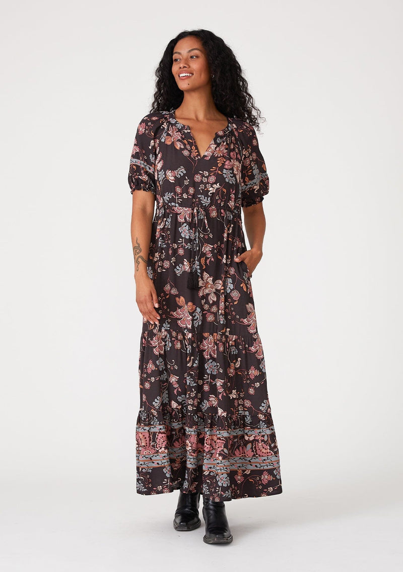 [Color: Brown/Dusty Lilac] A front facing image of a brunette model wearing a bohemian fall maxi dress in a brown and dusty purple floral print. With short puff sleeves, a v neckline, a long tiered skirt, side pockets, and a drawstring waist with tassel ties. 