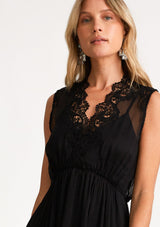 [Color: Black] A close up front facing image of a blonde model wearing a black bohemian holiday mid length special occasion dress. A sleeveless holiday dress designed in chiffon, with lace trim, a surplice v neckline, an empire waist, a half smocked bodice at the back, and an open back detail with single button closure. 