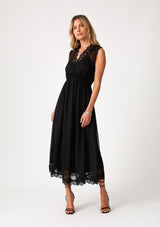 [Color: Black] A front facing image of a blonde model wearing a black bohemian holiday mid length special occasion dress. A sleeveless holiday dress designed in chiffon, with lace trim, a surplice v neckline, an empire waist, a half smocked bodice at the back, and an open back detail with single button closure. 