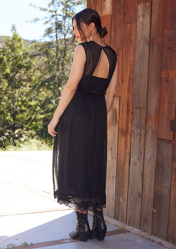 [Color: Black] A back facing image of a brunette model standing outside wearing a black bohemian holiday mid length special occasion dress. A sleeveless holiday dress designed in chiffon, with lace trim, a surplice v neckline, an empire waist, a half smocked bodice at the back, and an open back detail with single button closure.