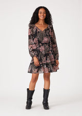 [Color: Black/Dusty Rose] A front facing image of a brunette model wearing a bohemian fall mini dress in a black and pink floral print. With voluminous long sleeves, a split v neckline with ties, a tiered mini skirt, a smocked elastic waist, and sheer mesh details. 