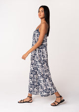 [Color: Navy/Natural] A side facing image of a brunette model wearing a sleeveless blue floral one piece jumpsuit. With adjustable spaghetti straps, a scoop neckline, a cropped wide leg, and a loose, relaxed fit. 
