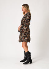 [Color: Black/Taupe] A side facing image of a blonde model wearing a bohemian fall mini dress in a bohemian brown print. With an empire waist, a deep v neckline, a relaxed flowy fit, and long bell sleeves with a split wrist detail. 