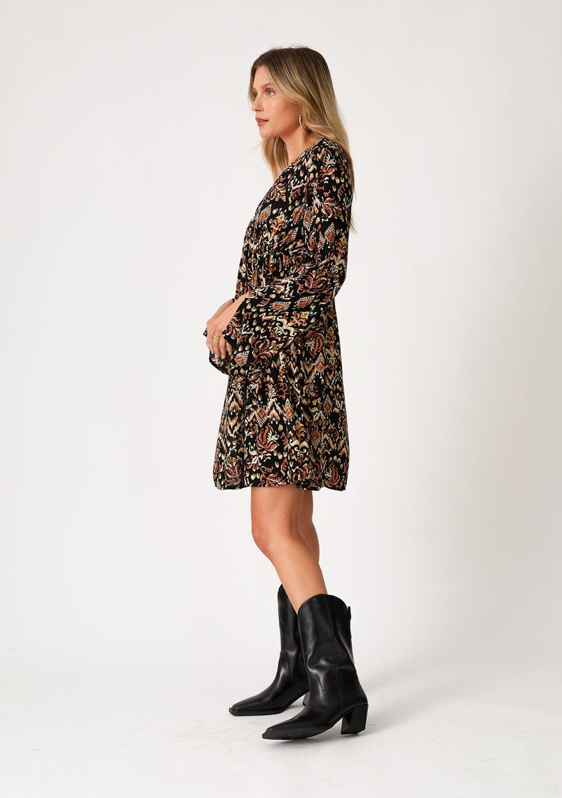 [Color: Black/Taupe] A side facing image of a blonde model wearing a bohemian fall mini dress in a bohemian brown print. With an empire waist, a deep v neckline, a relaxed flowy fit, and long bell sleeves with a split wrist detail. 