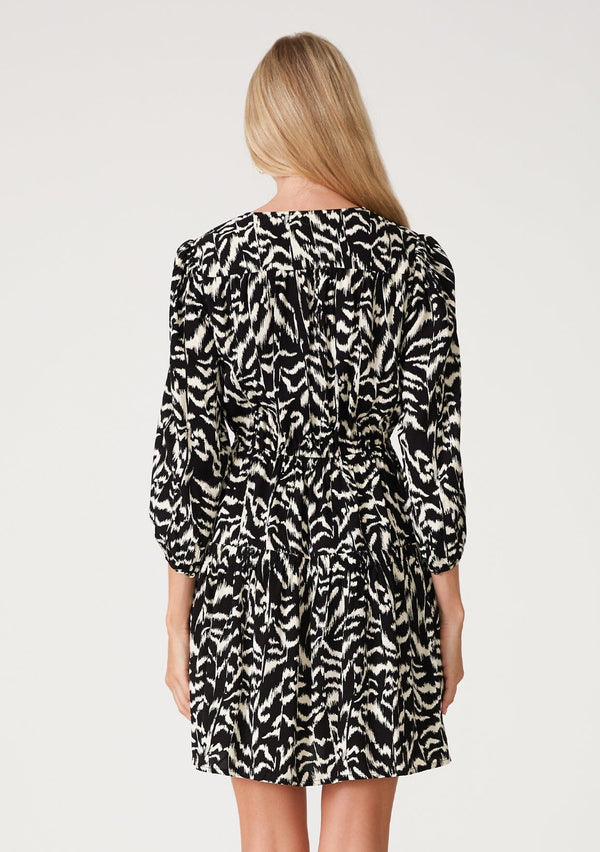 [Color: Black/Natural] A back facing image of a blonde model wearing a casual resort mini dress in an abstract bohemian black and off white print. With three quarter length sleeves with a single button closure, a surplice v neckline, and a tiered skirt. 