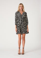 [Color: Black/Natural] A full body front facing image of a blonde model wearing a casual resort mini dress in an abstract bohemian black and off white print. With three quarter length sleeves with a single button closure, a surplice v neckline, and a tiered skirt. 