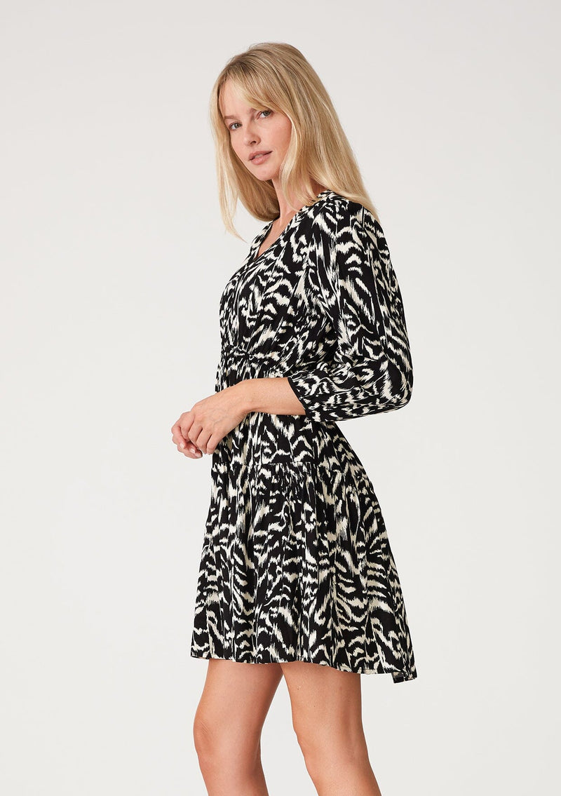 [Color: Black/Natural] A side facing image of a blonde model wearing a casual resort mini dress in an abstract bohemian black and off white print. With three quarter length sleeves with a single button closure, a surplice v neckline, and a tiered skirt. 