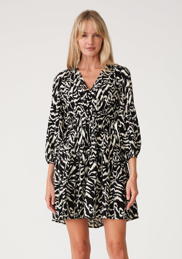 [Color: Black/Natural] A front facing image of a blonde model wearing a casual resort mini dress in an abstract bohemian black and off white print. With three quarter length sleeves with a single button closure, a surplice v neckline, and a tiered skirt. 