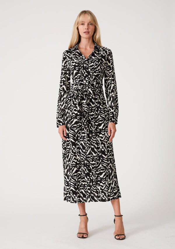 [Color: Black/Natural] A front facing image of a blonde model wearing a bohemian resort maxi shirt dress in a black and off white abstract print. With a self covered button front, long sleeves, a collared neckline, side slits, and an attached waist tie. 