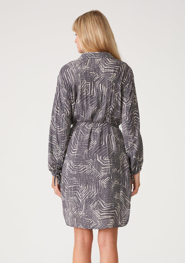 [Color: Grey/Cream] A back facing image of a blonde model wearing a relaxed fit mini shirt dress in an abstract grey print. With long sleeves, a gathered drawstring sleeve detail, tie wrist cuffs, a high low hemline, a self covered button front, side pockets, a collared neckline, and a tie waist belt. 