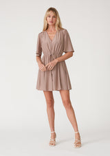 [Color: Mocha] A full body front facing image of a blonde model wearing a mocha brown bohemian resort mini dress. With short flutter sleeves, a v neckline, an adjustable drawstring tie waist, and a mini pom trim. 