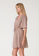 [Color: Mocha] A side facing image of a blonde model wearing a mocha brown bohemian resort mini dress. With short flutter sleeves, a v neckline, an adjustable drawstring tie waist, and a mini pom trim. 