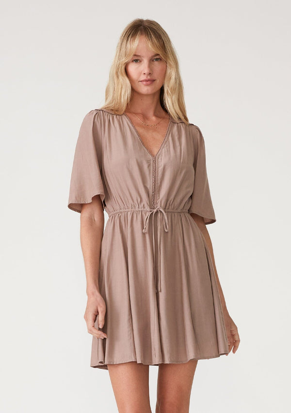 [Color: Mocha] A front facing image of a blonde model wearing a mocha brown bohemian resort mini dress. With short flutter sleeves, a v neckline, an adjustable drawstring tie waist, and a mini pom trim. 
