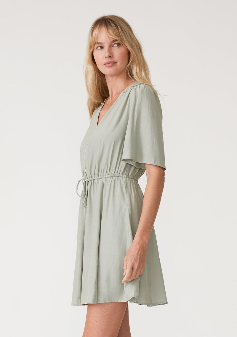 [Color: Pistachio] A side facing image of a blonde model wearing a light green bohemian resort mini dress. With short flutter sleeves, a v neckline, an adjustable drawstring tie waist, and a mini pom trim. 