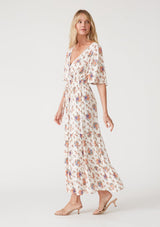 [Color: Natural/Rust] A side facing image of a blonde model wearing a classic bohemian maxi dress in an off white and pink floral print. With short flutter sleeves, a surplice v neckline, a smocked elastic waist, a drawstring tie waist, and a long flowy tiered skirt. 