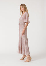 [Color: Ivory/Purple] A side facing image of a blonde model wearing a classic bohemian maxi dress in an off white and purple ditsy floral print. With short flutter sleeves, a surplice v neckline, a smocked elastic waist, a drawstring tie waist, and a long flowy tiered skirt.