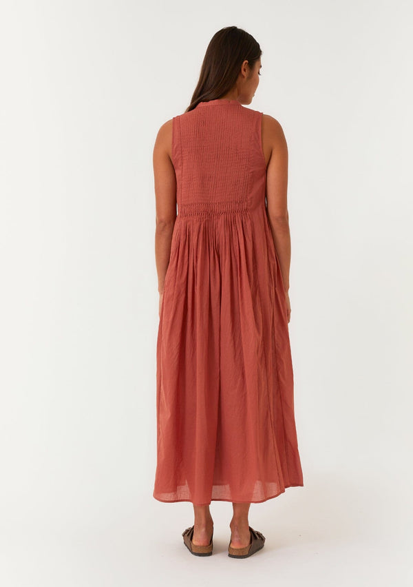 [Color: Rust] A back facing image of a brunette model wearing a lightweight bohemian sleeveless maxi tent dress in rust red. With a v neckline, pleated details, and side pockets. 