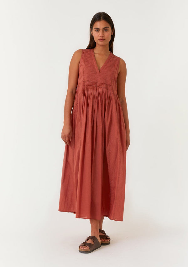 [Color: Rust] A front facing image of a brunette model wearing a lightweight bohemian sleeveless maxi tent dress in rust red. With a v neckline, pleated details, and side pockets. 