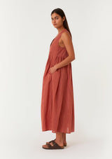 [Color: Rust] A side facing image of a brunette model wearing a lightweight bohemian sleeveless maxi tent dress in rust red. With a v neckline, pleated details, and side pockets. 