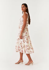 [Color: Natural/Dusty Wine] A side facing image of a brunette model wearing a sleeveless bohemian mid length dress in an off white and pink floral print. With a ruffle trimmed scoop neckline, a self covered button front top, a tiered skirt, lace trim, pintuck details, and a drawstring waist with ties. 