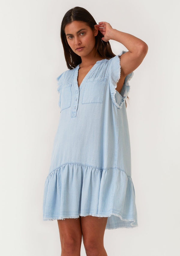 [Color: Bleach Wash] A front facing image of a brunette model wearing a denim blue bohemian spring mini dress crafted from Tencel. With short flutter sleeves, a v neckline, a tiered mini skirt, front patch pockets, a self covered button front, a raw hemline, and pleated details. 