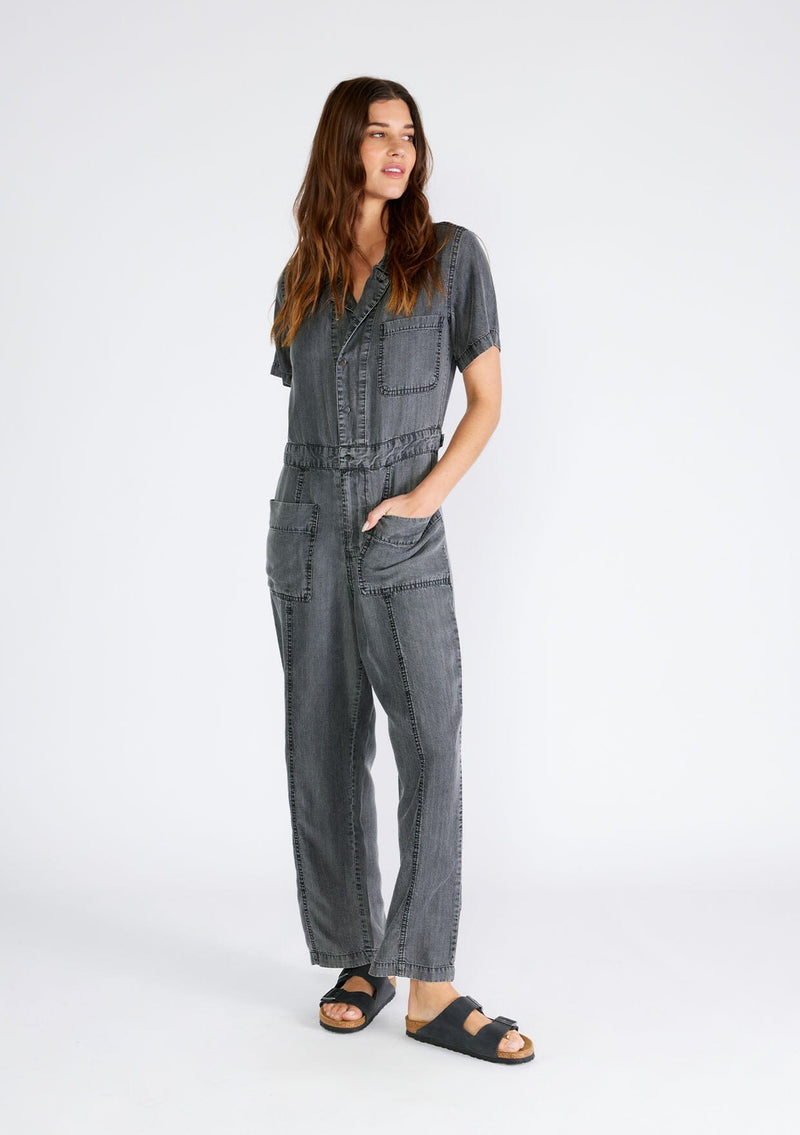 [Color: Ash Grey Wash] A full body front facing image of a brunette model wearing a utility style jumpsuit in an ash grey wash. Designed in Tencel, with short sleeves, a collared neckline, a long straight leg, a button front top, a zip fly closure, and front and back patch pockets. 