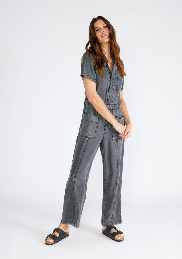 [Color: Ash Grey Wash] A front facing image of a brunette model wearing a utility style jumpsuit in an ash grey wash. Designed in Tencel, with short sleeves, a collared neckline, a long straight leg, a button front top, a zip fly closure, and front and back patch pockets. 