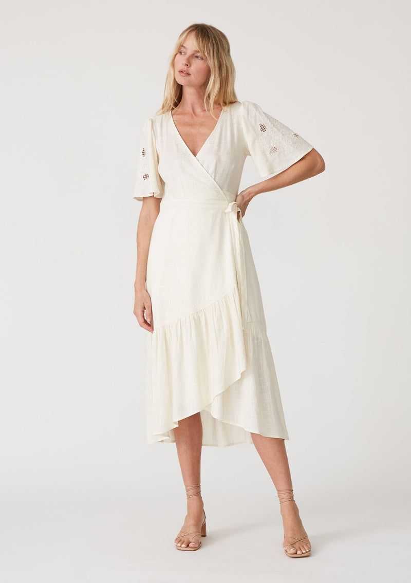 [Color: Vanilla] A front facing image of a blonde model wearing an off white bohemian mid length wrap dress. With short flutter sleeves, embroidered detail, a deep v neckline, a side tie waist closure, and a flowy tiered skirt. 