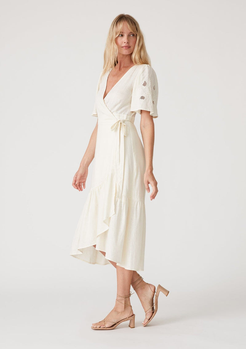 [Color: Vanilla] A side facing image of a blonde model wearing an off white bohemian mid length wrap dress. With short flutter sleeves, embroidered detail, a deep v neckline, a side tie waist closure, and a flowy tiered skirt. 